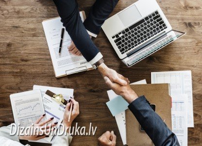 Деловые партнеры пожимают руки над столом с ноутбуком и документами, обсуждая визитные карточки