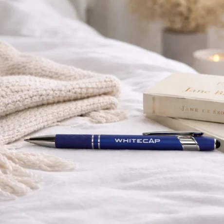 Синяя металлическая ручка с гравировкой WHITECAP на кровати рядом с книгами и пледом