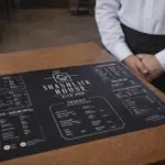 Black A3 flat custom menu on a wooden restaurant table with waiter in the background