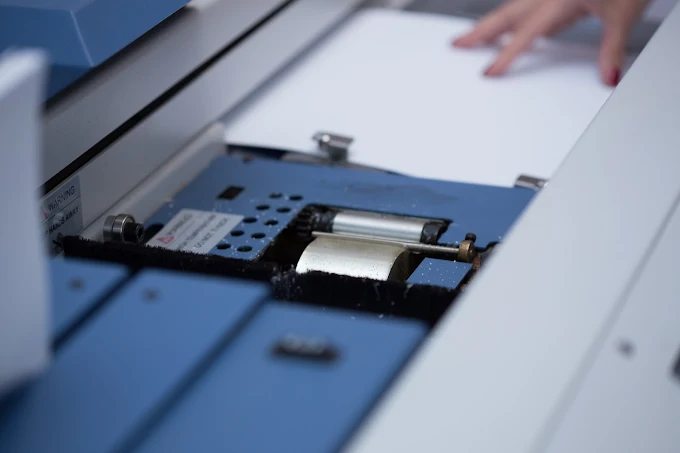 A hand places a white sheet on a blue industrial machine beside metal rollers