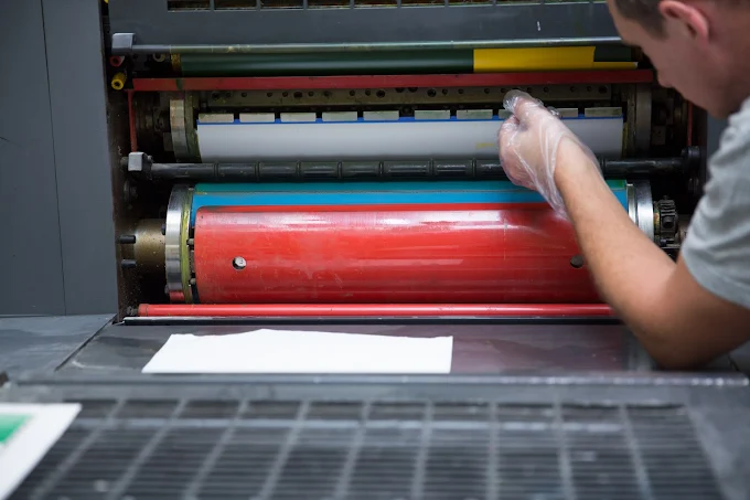 A gloved person adjusts the colored rollers of a printing press, with a white sheet in front