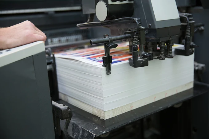 Printing machine processes a large stack of white paper sheets, with a hand visible beside it